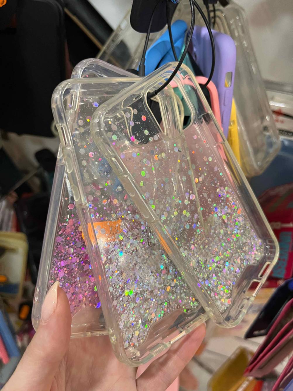 Drei am Verkaufsstand hängende, getestete iPhone-12-Mini-Hüllen mit Glitzereffekt werden von einer Damenhand inspiziert.