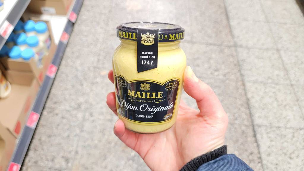 Maille-Senf-Test: Eine Hand hält ein Glas original Dijon-Senf vor dem Boden eines Supermarktes.