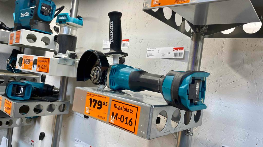 Ansicht von schräg hinten auf einen Makita-Akku-Winkelschleifer im Test. Das türkis-schwarze Modell liegt ohne Akku in einem Verkaufsbereich neben weiteren Makita-Werkzeugen.