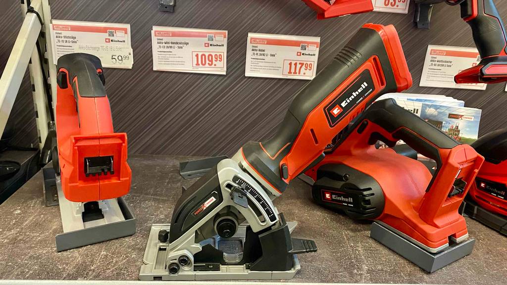 Inmitten verschiedener Einhell-Geräte auf einem Ausstellungsregal steht eine Mini-Handkreissäge im Test: Das grau-rote Einhell-Modell präsentiert sich von der Seite inklusive Parallelanschlag.