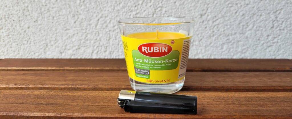 Eine gelbe getestete Anti-Mücken-Kerze steht hinter einem liegenden Feuerzeug auf einer Holzfläche.