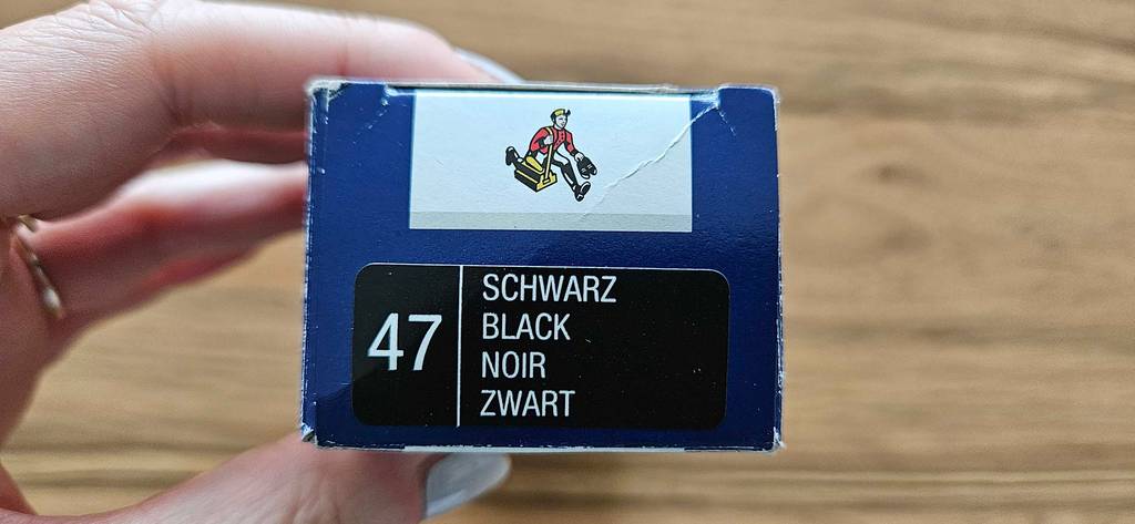 Hand hält eine blaue Verpackung einer getesteten Schuhcreme, mit Fokus auf die Produktangabe „Schwarz“, vor beigefarbenem Hintergrund.