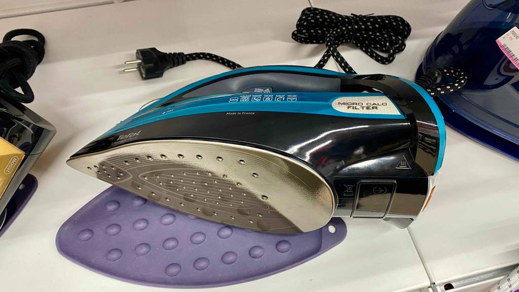 Tefal-Bügeleisen im Test: ein türkis-schwarzes Modell liegt mit Fokus auf die gelöcherte Edelstahl-Unterseite auf einem weißen Regal zwischen anderen Bügeleisen.