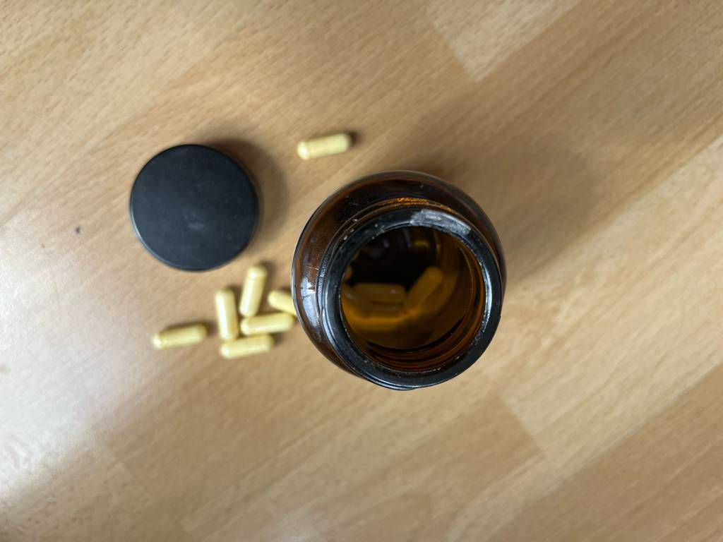 Vitamin-B6-Test: Blick von oben auf eine blickdichte geöffnete Glasdose mit schwarzem Drehverschluss, die auf einer weißen Holzfläche neben hellen Kapseln steht.