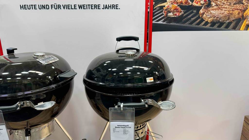 Zwei runde, getestete Weber-Holzkohlegrills inklusive Standfüßen stehen nebeneinander in einem Baumarkt.