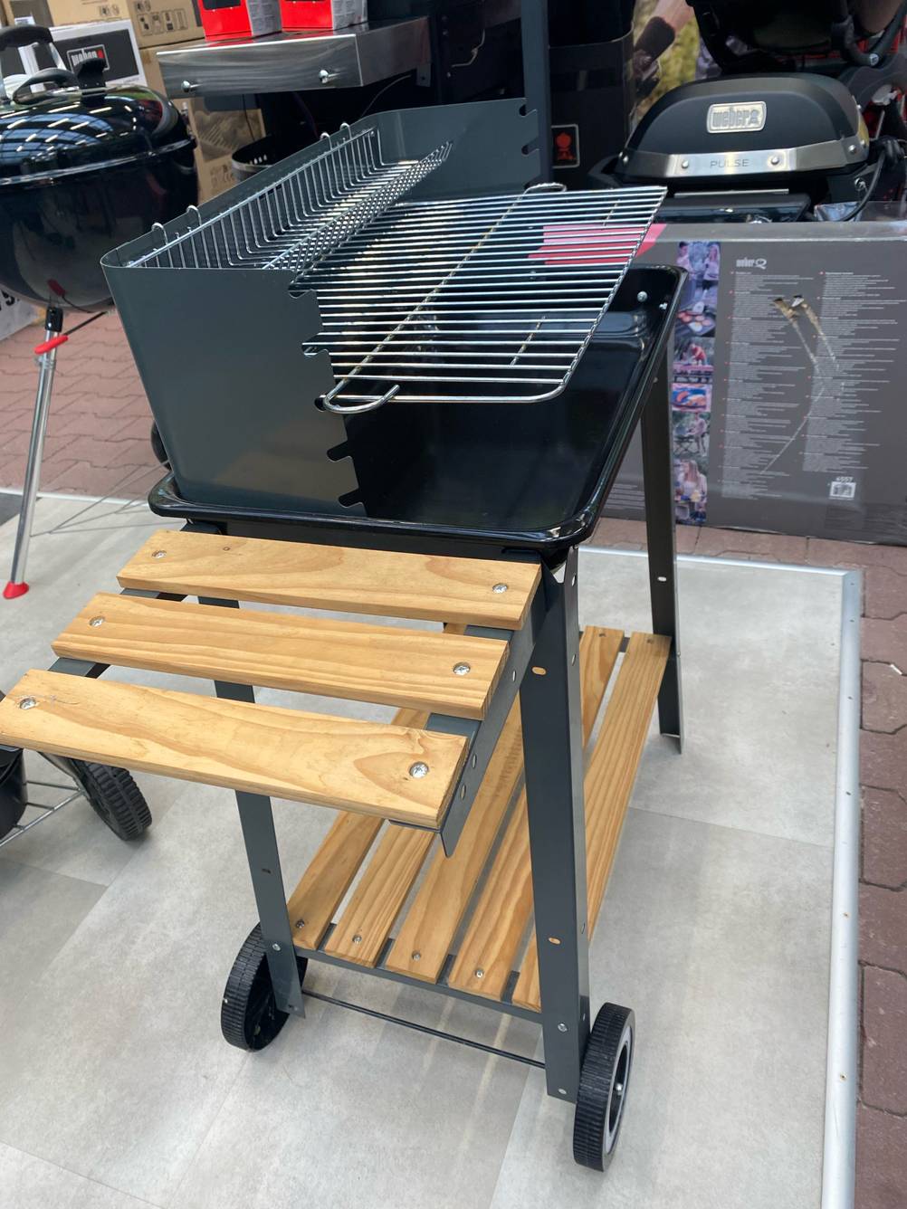 Seitenansicht eines fahrbaren, getesteten Grillwagens: eine Grillfläche ist höhenverstellbar und zwei Holzablagen im Fußbereich und seitlich neben dem Grill.