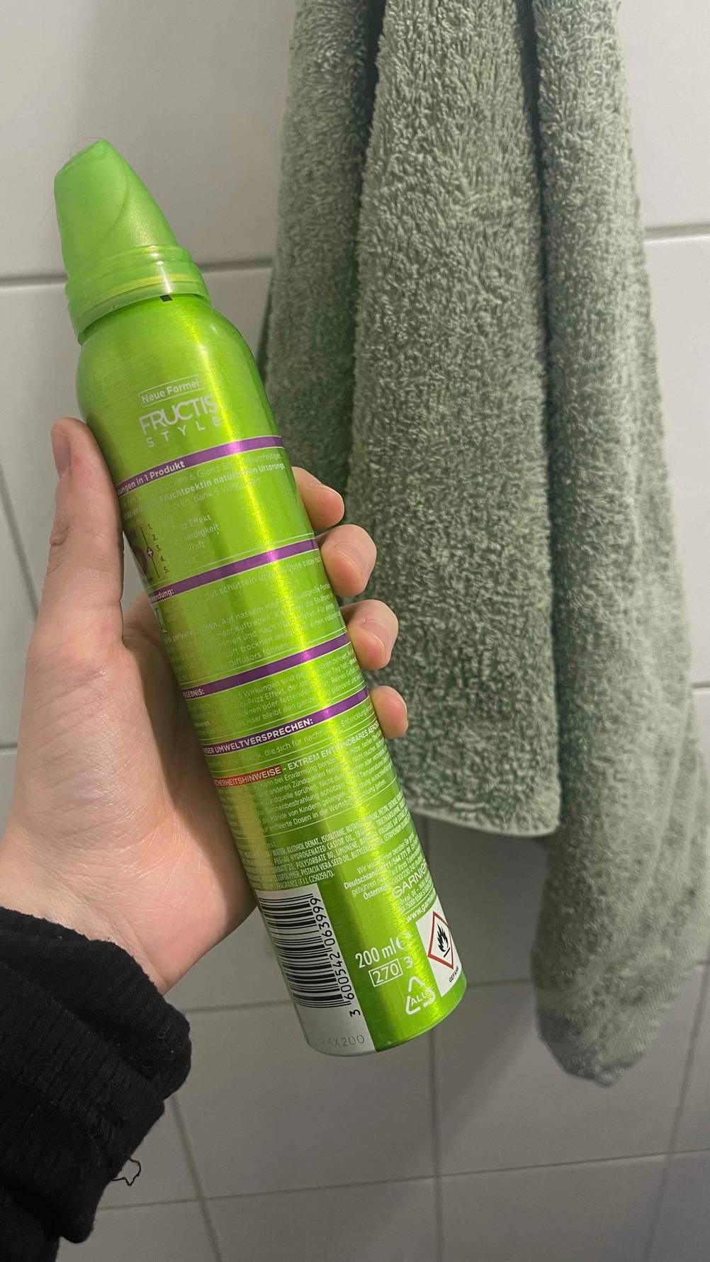 Eine Hand hält den giftgrünen 200-ml-Spender inklusive Produktbeschreibung des Schaumfestigers für Locken im Test vor einem olivgrünen Handtuch.