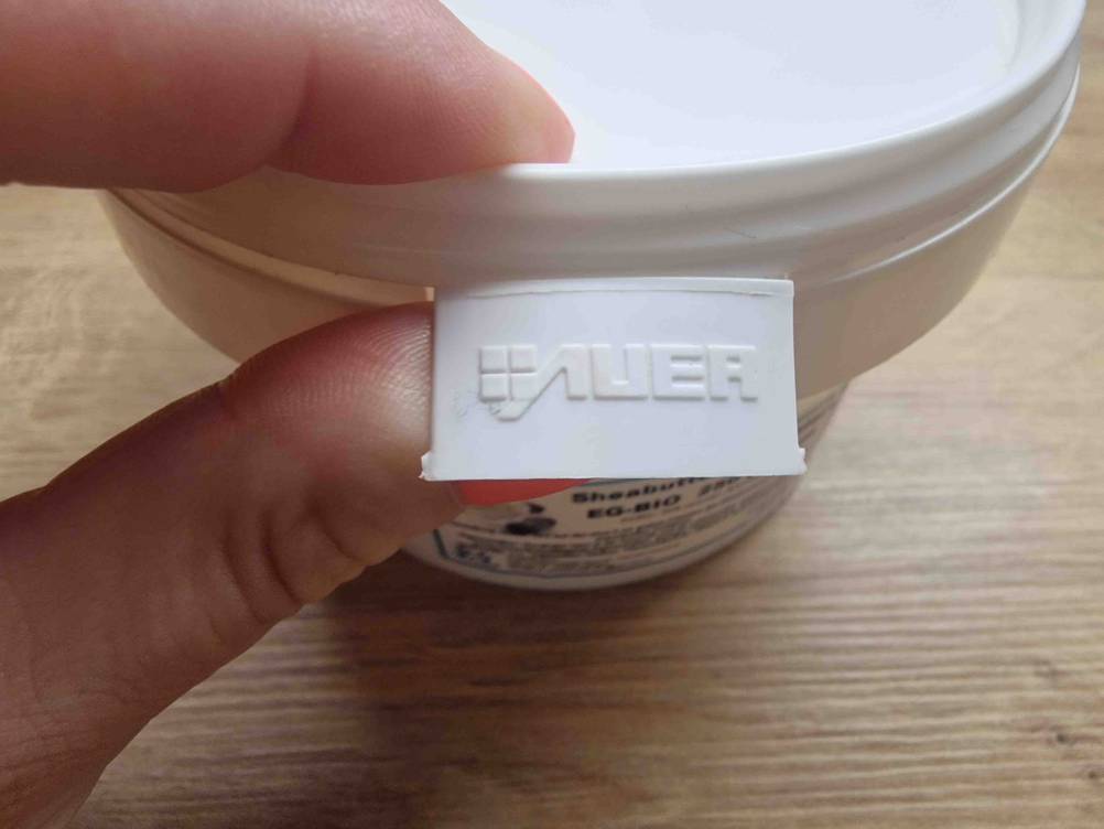 Getestet: Detailansicht eines weißen Sheabutter-Tiegels, mit Fingern, die die Öffnungs-Lasche mit der Aufschrift Auer halten.