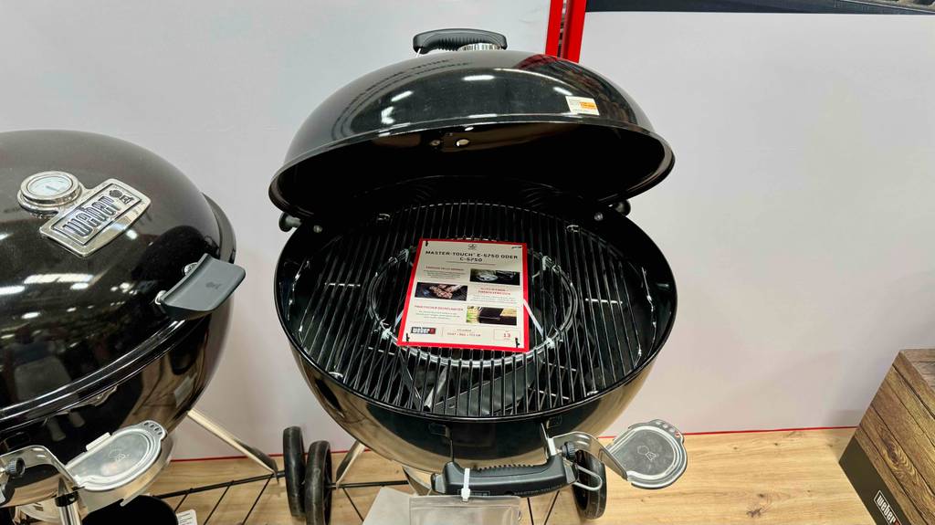 Der runde Weber-Holzkohlegrill im Test steht mit geöffnetem Deckel, der Fokus liegt auf dem Grillgut-Rost, neben einem anderen runden Standgrill von Weber in einem Baumarkt.