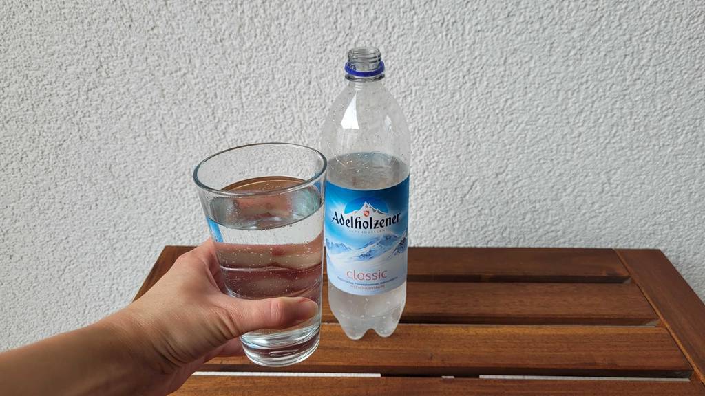 Adelholzener-Wasser-Test: Eine Hand greift nach einem Glas Mineralwasser, das aus der Kunststoffflasche stammt.