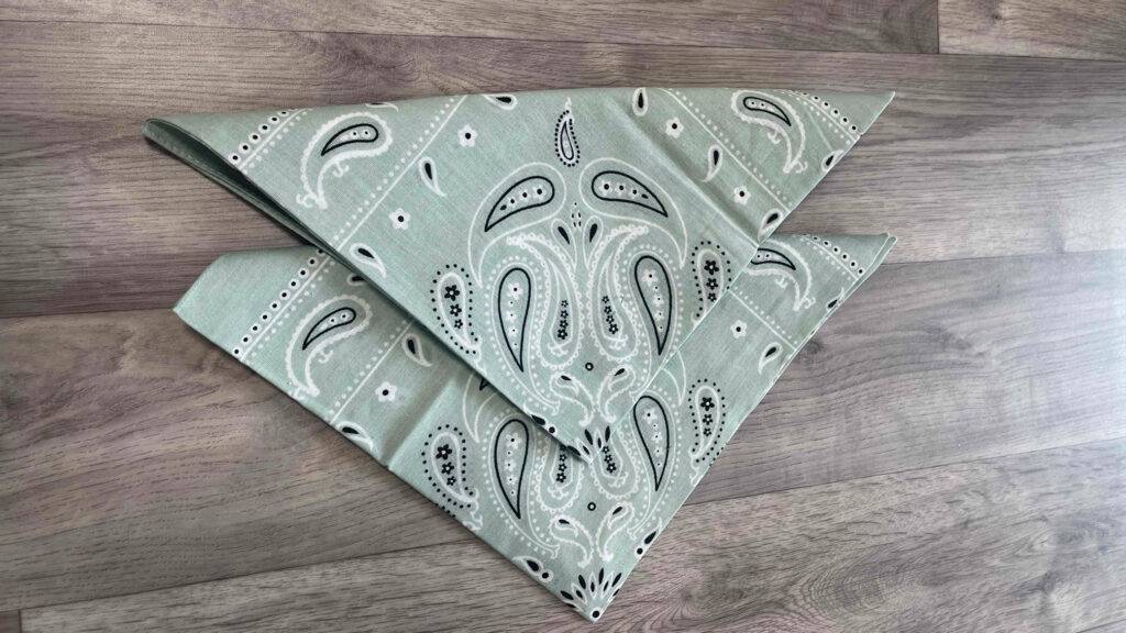 Zwei im Dreieck gefaltete, getestete Bandana Kopftücher liegen nebeneinander auf einer Holzoberfläche.