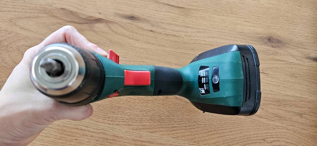 Eine Hand hält einen grünen getesteten Bosch-Akkuschrauber, mit Fokus auf die rote Einschalttaste und die Batteriestandanzeige, vor einer Holzoberfläche.