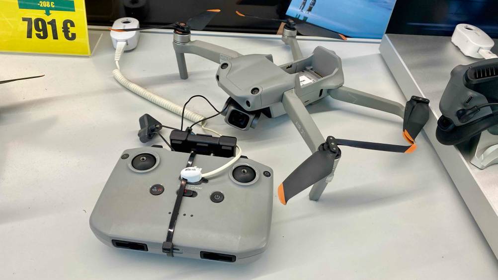 Eine hellgraue DJI-Drohne im Test liegt inklusive Steuerung auf einer weißen Verkaufsfläche neben einem gelben Preisschild.