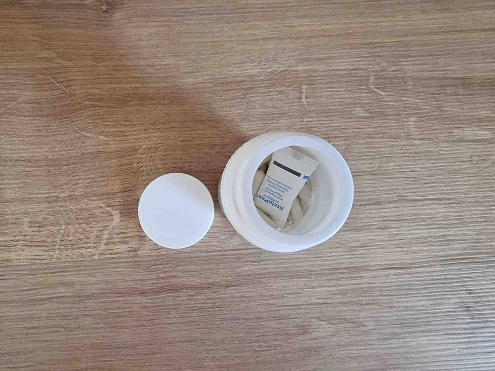 Eisenpräparat-Test: weiße Kapseln in einer weißen geöffneten Kunststoff-Dose inklusive Deckel auf einer Holzfläche.