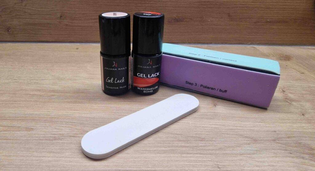 Vor zwei schwarzen Behältern mit Gel-Nagellacken im Test liegt ein Polier-Pad, inklusive Verpackung, auf einer Holzfläche.