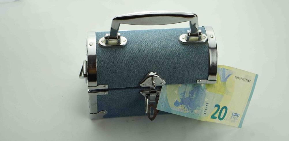 Gentestes Geldversteck mit Metall-Schlossvorrichtung an einem mit Jeansstoff verkleideten Köfferchen inklusive 20-Euro-Schein vor weißem Hintergrund.