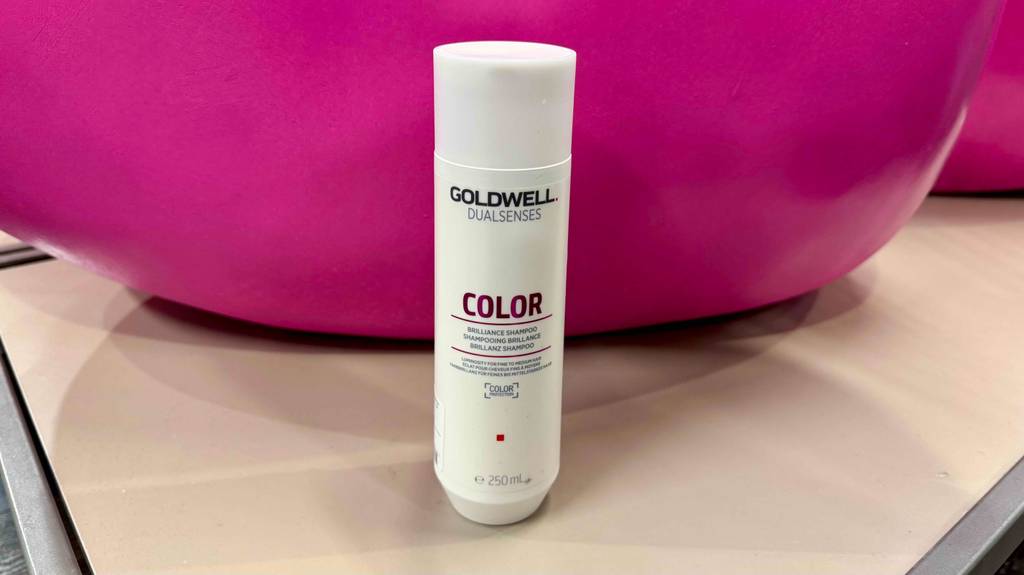Ein getestetes Goldwell-Shampoo in einer weißen Kunststoffflasche steht auf einer weißen Fläche vor einem pinkfarbenen Gegenstand.