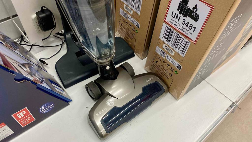 Grundig-Akku-Staubsauger im Test: ein silberfarbener Stielstaubsauger, mit Fokus auf den Bürstenaufsatz und den transparenten Staubfangbehälter lehnt neben seiner braunen Verpackung auf einem weißen Verkaufsregal.
