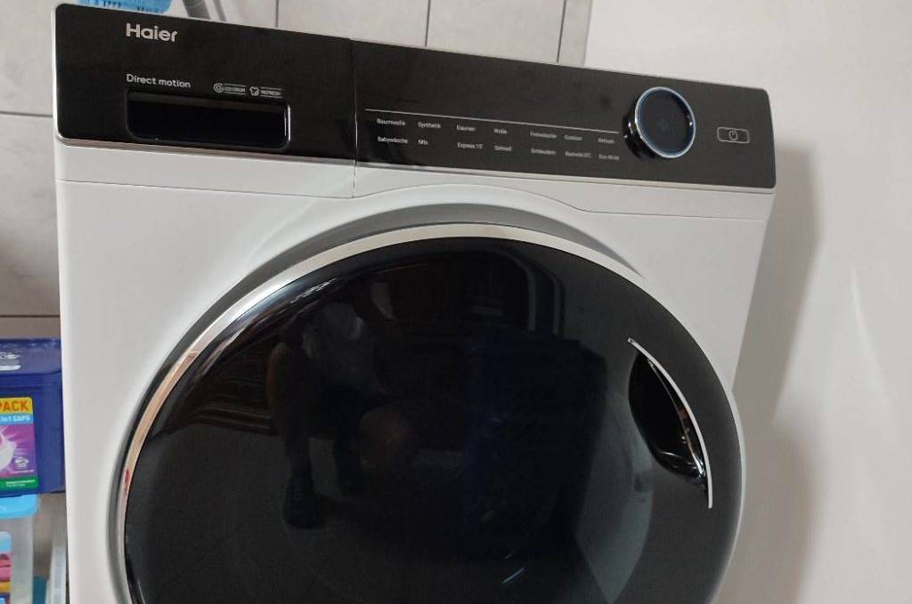 Weiße Haier-Waschmaschine im Test inklusive schwarzer Oblo-Abdeckung steht an Wasser angeschlossen an einer weißen Wand.