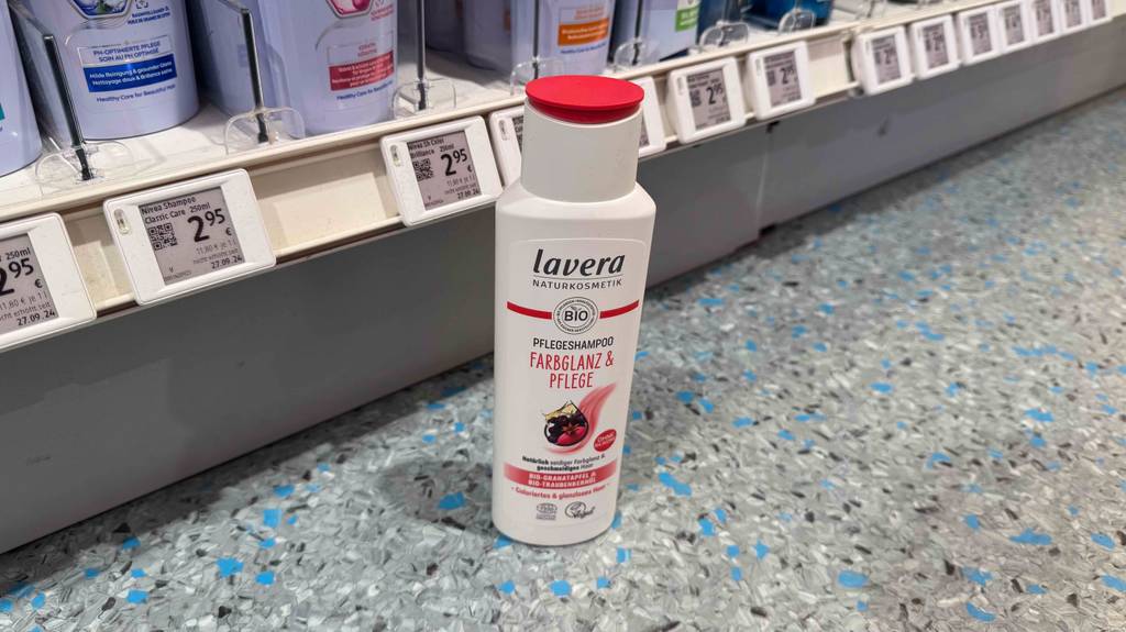 Eine weiße Kunststoffflasche mit getestetem Lavera-Shampoo steht auf einem Linoleumboden vor einem Verkaufsregal mit Preisschildern.