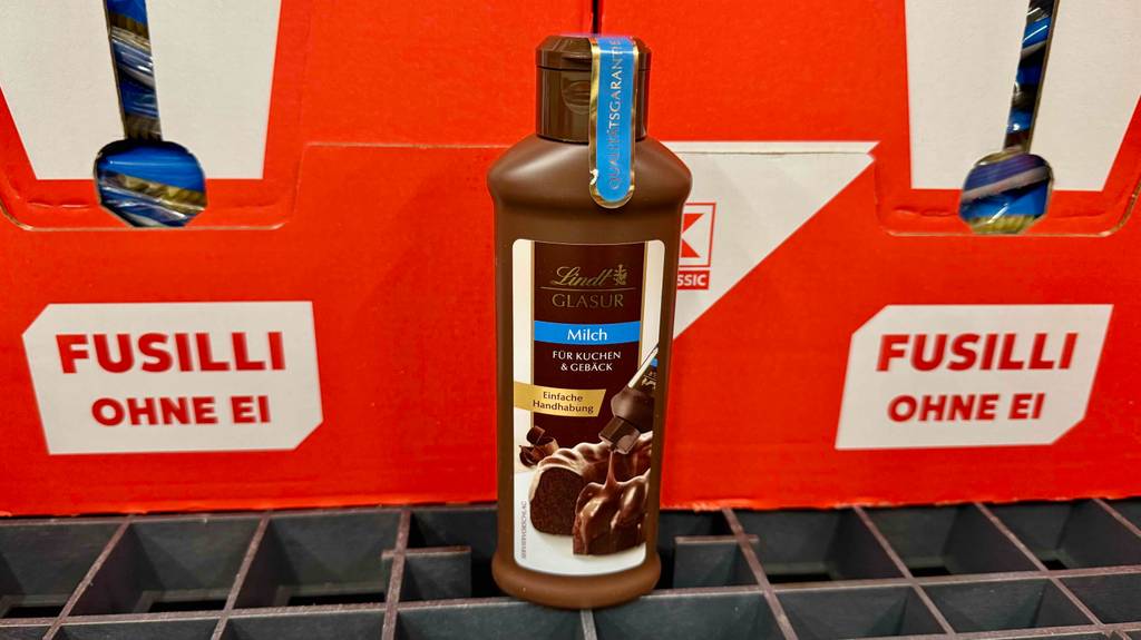 Eine braune Quetschflasche mit getesteter Kuchenglasur von Lindt steht vor Kartons mit Nudeln.