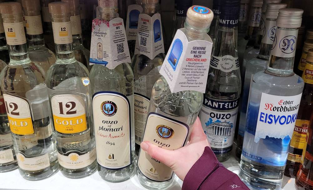 Eine Hand greift in einem Flaschenregal nach einer transparenten Glasflasche inklusive Ouzo im Test, der Marke Plomari, mit Korkverschluss.