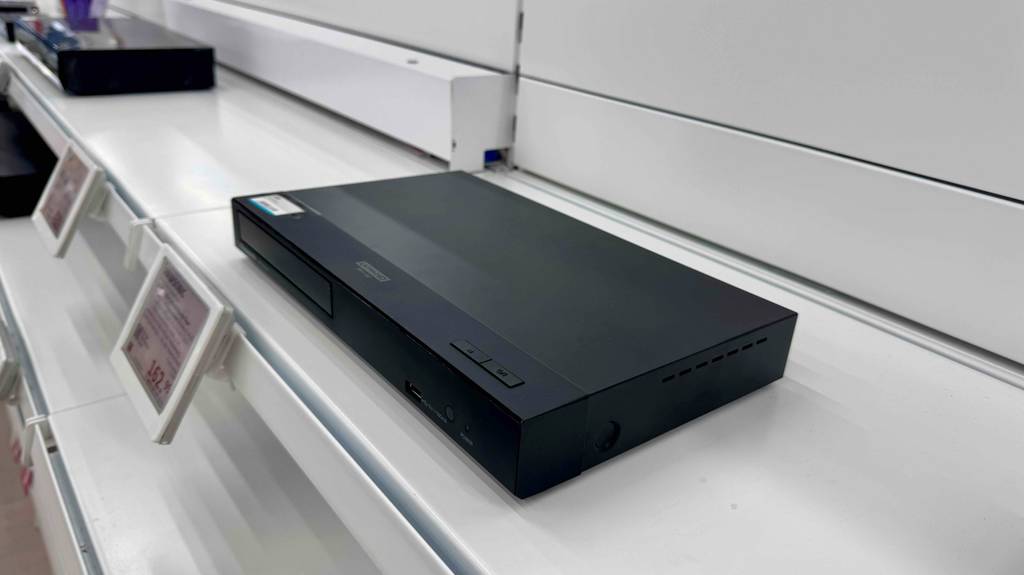 Im Test: Der HD-Receiver der Marke Panasonic, von der Seite betrachtet, steht auf einem weißen Regal mit Preisschildern.