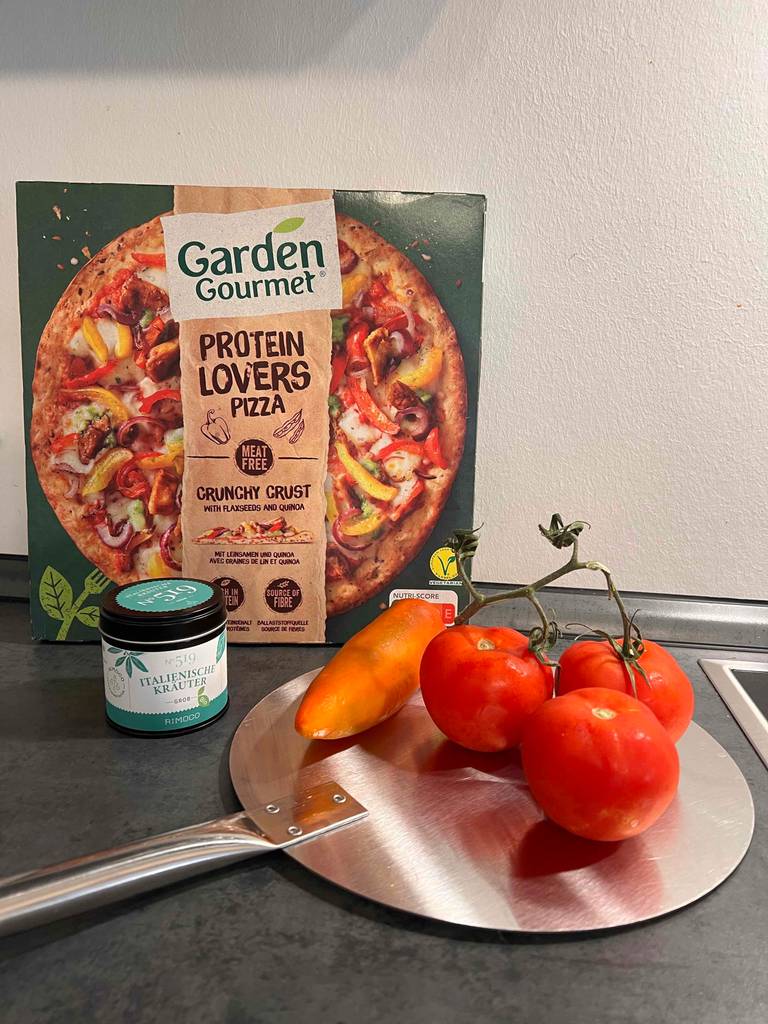 Eine getestete Pizzaschaufel trägt drei Tomaten und eine Paprika und liegt vor einer Fertigpizza.