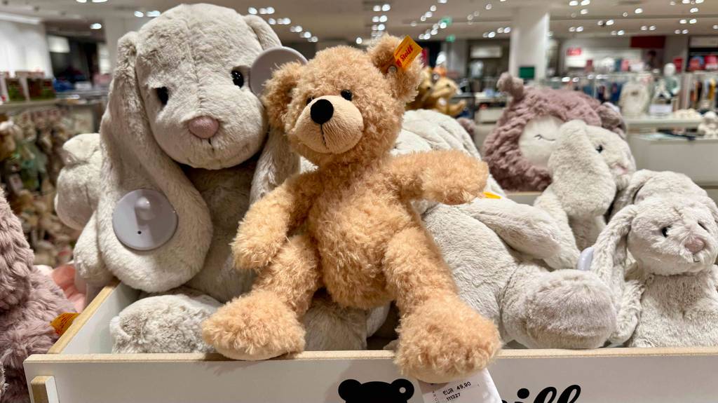 Mehrere Stofftiere, darunter ein getesteter Steiff-Teddy, sitzen nebeneinander in einem weißen Plüschtier-Behälter im Geschäft.
