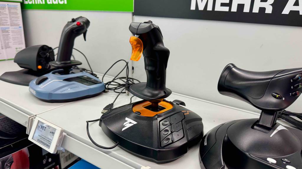 Thrustmaster-Joysticks im Test: Das schwarze Modell mit orangefarbenen Highlights steht zwischen weiteren Joysticks auf einem weißen Ausstellungsregal.