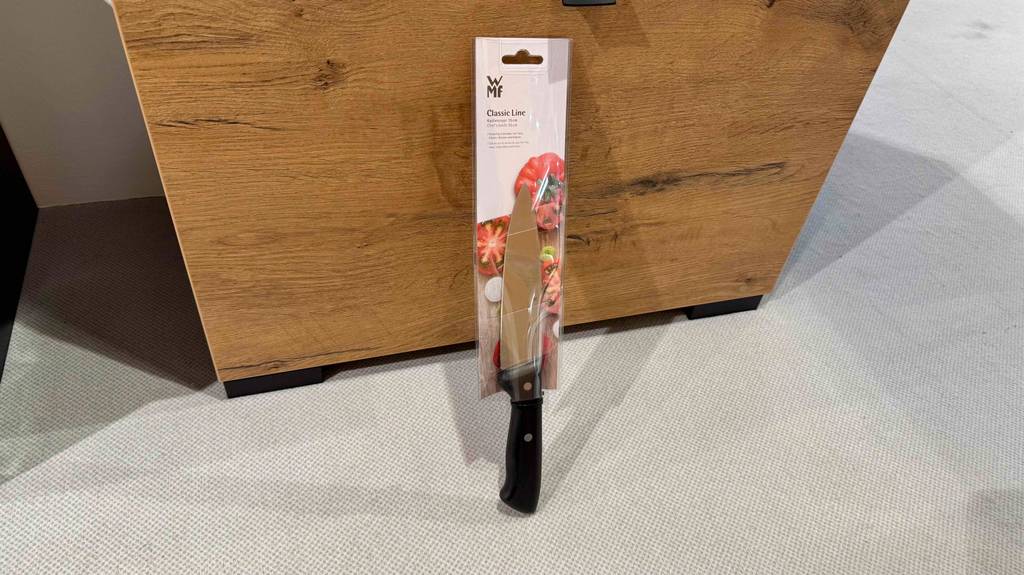 Getestet: Ein WMF-Kochmesser mit schwarzem Griff lehnt mit der Verpackung an einer Holzkiste.