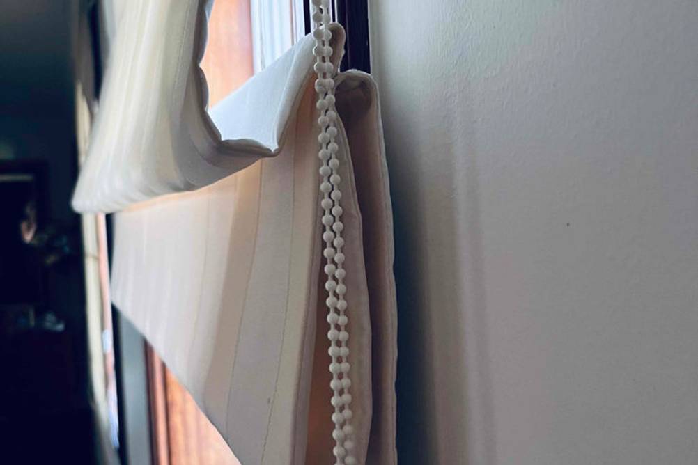 Raffrollo im Test: seitliche Nahaufnahme des Kordelzugs eines hellen Raffrollos leicht hochgerafft vor einem Fenster.