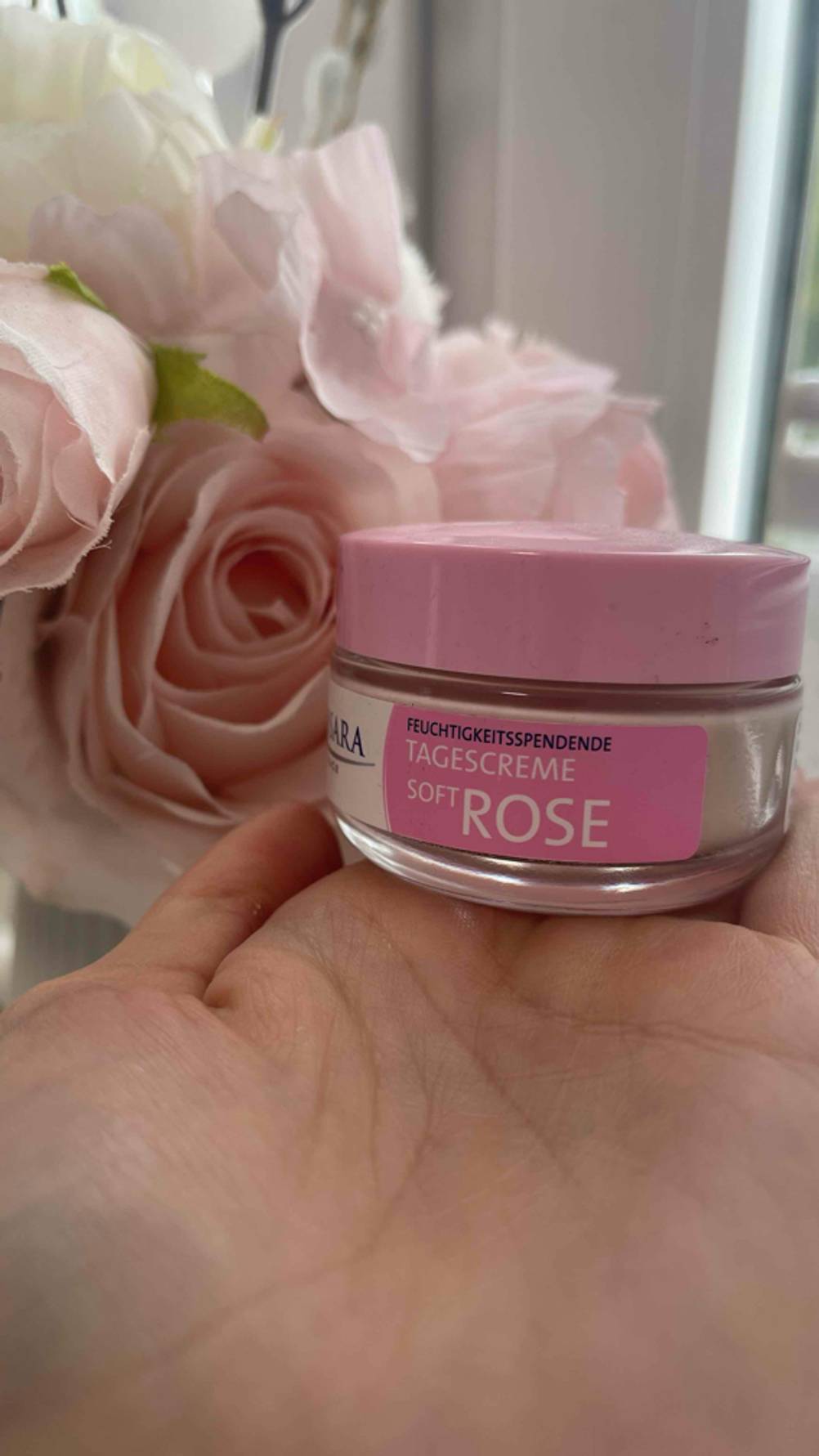 Im Test: Die Rosencreme im Tiegel liegt auf einer Handfläche vor rosafarbenen Rosen.