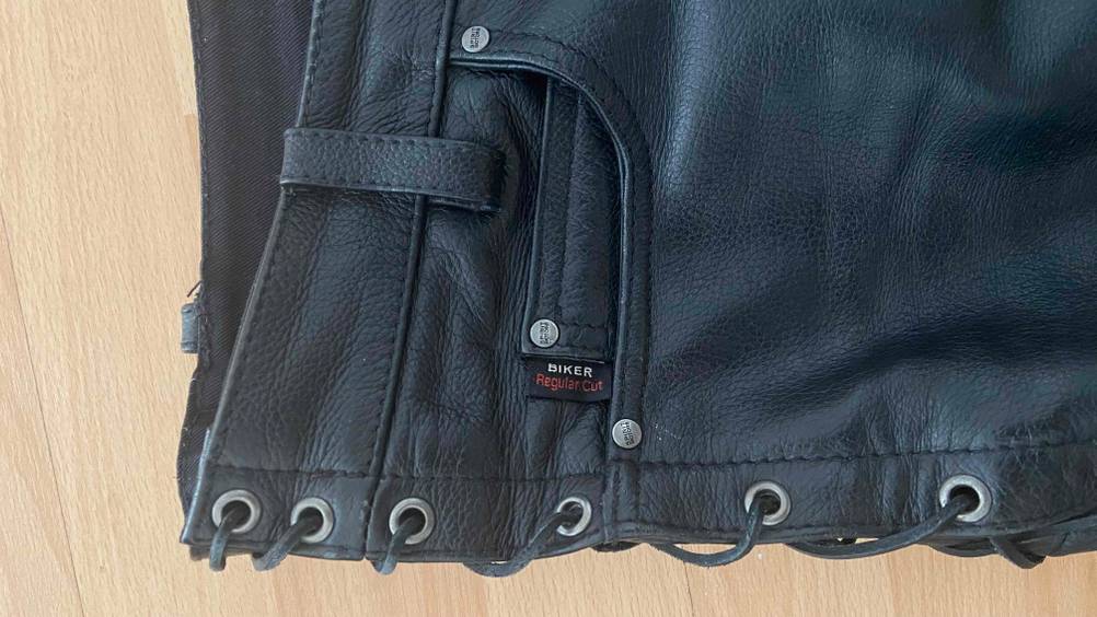 Lederhose im Test: Detailansicht der oberen, seitlichen Schnürung und Taschenöffnung einer schwarzen Lederhose auf einem Parkettboden.
