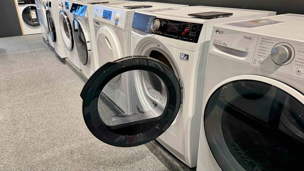 Seitenansicht eines getesteten AEG-Trockners mit geöffneter Rechtsanschlag-Tür, wie er zwischen weiteren Trocknern auf einer Ausstellungsfläche steht.