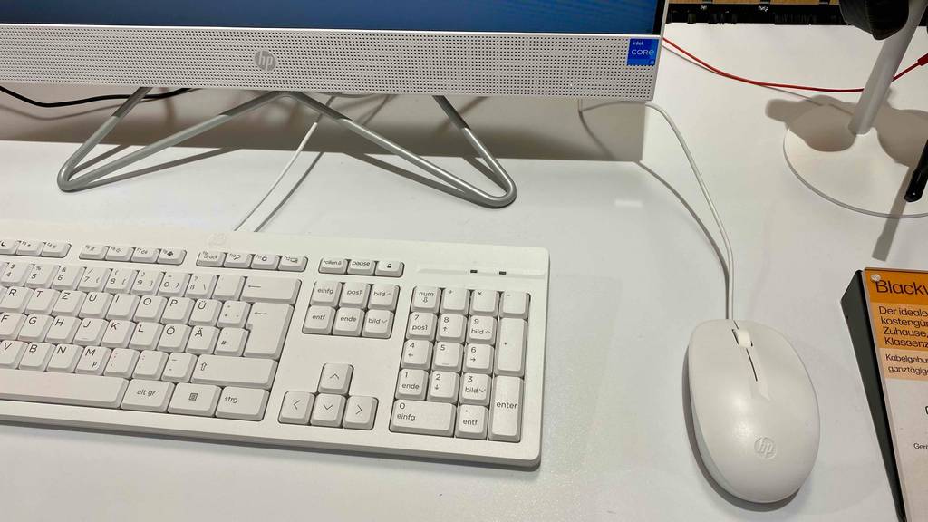 Detailaufnahme der weißen Tastatur inklusive Maus mit Kabelanschluss vor einem HP-Bildschirm eines getesteten weißen All-in-One-PCs im Verkauf.
