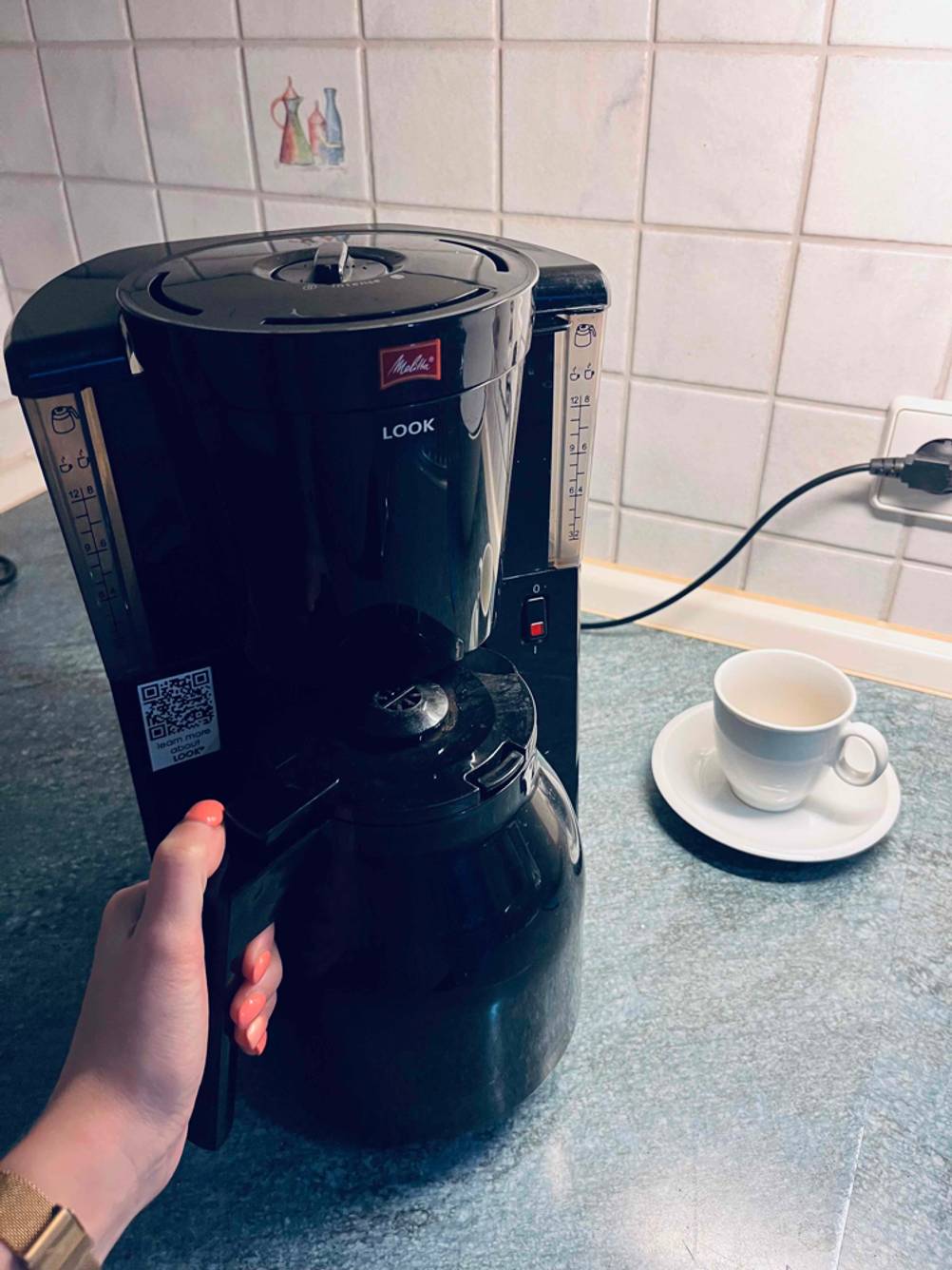 Hand greift nach schwarzer Kunststoff-Kanne einer Melitta Kaffeemaschine mit Direktbrühsystem, die auf einer hellgrauen Fläche neben einer weißen Kaffeetasse steht.