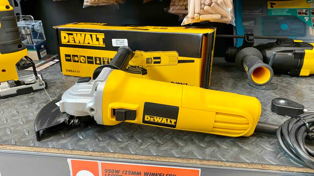 Seitenansicht auf einen gelben DeWalt Winkelschleifer im Test, der auf einem grauen Metallregal im Verkaufsbereich vor seiner Originalverpackung und weiteren Arbeitsgeräten liegt.