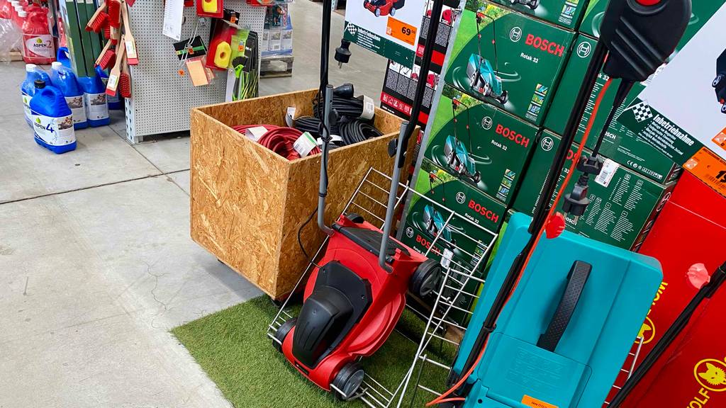 Einhell-Elektro-Rasenmäher-Test: ein rot-schwarzes Modell lehnt vor einer Verpackung mit einem Bosch-Rasenmäher im Verkaufsbereich.