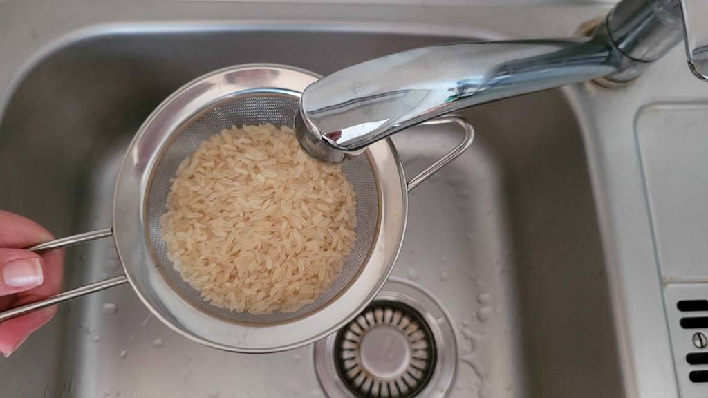 Küchensieb-Test: Hand hält ein Edelstahl-Küchensieb mit Vollkornreis unter einen Wasserhahn einer Küchenspüle.