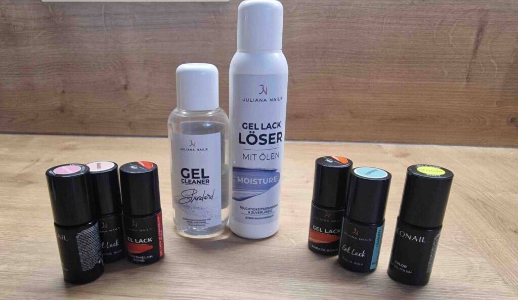 Zwischen mehreren schwarzen Behältern mit Gel-Nagellacken im Test in unterschiedlichen Farben stehen ein Flasche Gel-Lack-Löser und ein Gel-Lack Cleaner im weißen Design auf einer Holzfläche.