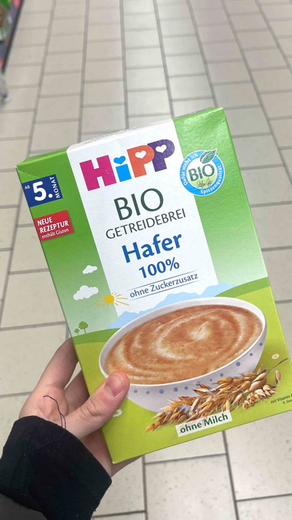 Getreidebrei-Test: Eine grüne Packung Haferbrei von Hipp wird von einer Hand über einem weißen Fliesenboden gehalten.