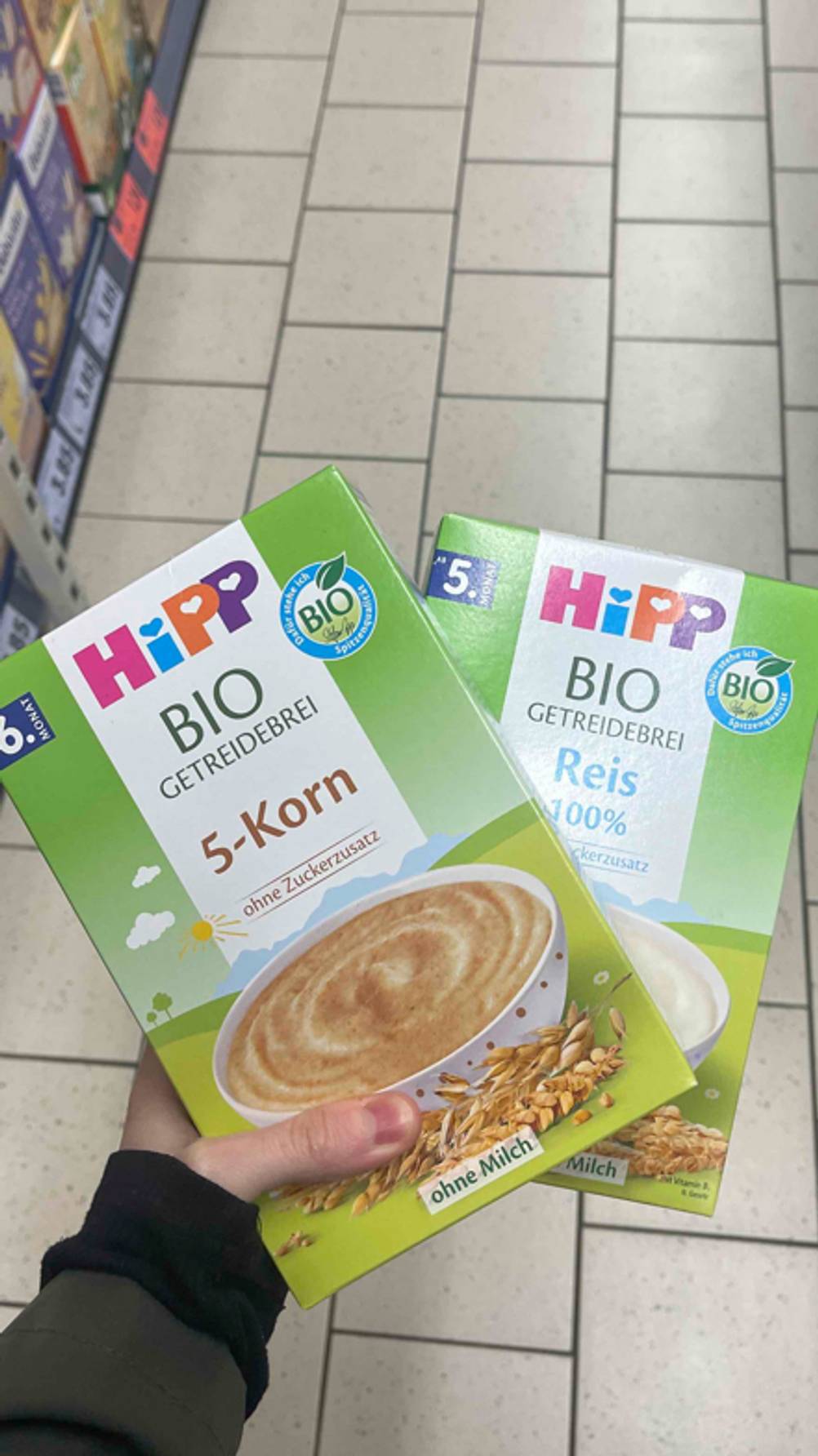 Eine Hand hält zwei getestete Getreidebrei-Packungen mit verschiedenen Geschmacksrichtungen über einem weißen Fliesenboden im Geschäft.