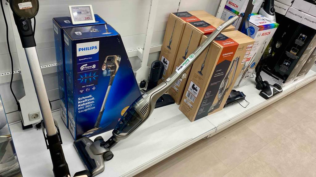 Grundig-Akku-Staubsauger-Test: ein silberfarbenes Modell lehnt an seiner braunen Verpackung vor einem Philips-Modell auf einem weißen Verkaufsregal.