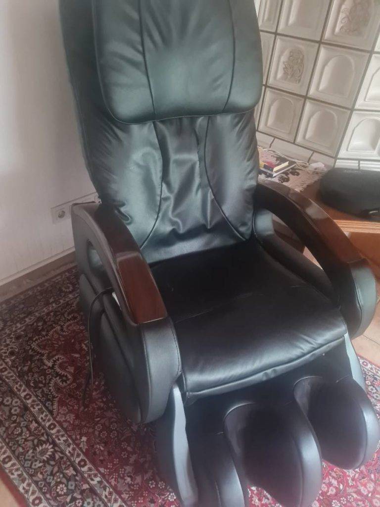 Getesteter Massagesessel im Leder-Design mit Holz-Armlehnen steht in einem Wohnbereich auf gemustertem Teppich.