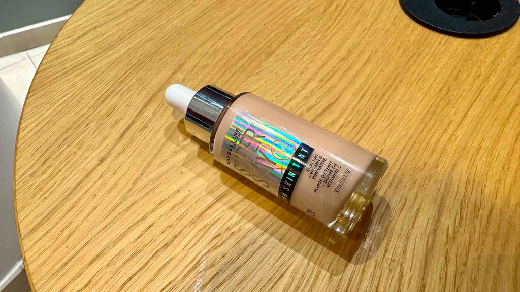 Ein getestetes Maybelline-Make-Up in der Flasche inklusive Pipette liegt auf einem Holztisch.