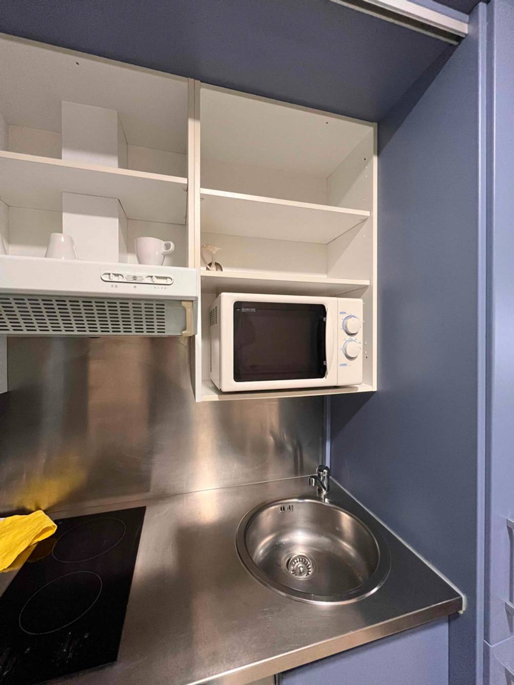 Miniküchen im Test: Über der in einer Aluminiumarbeitsplatte eingelassenen Aluminium-Spüle hängt ein weißer offener Schrank inklusive Mikrowelle.