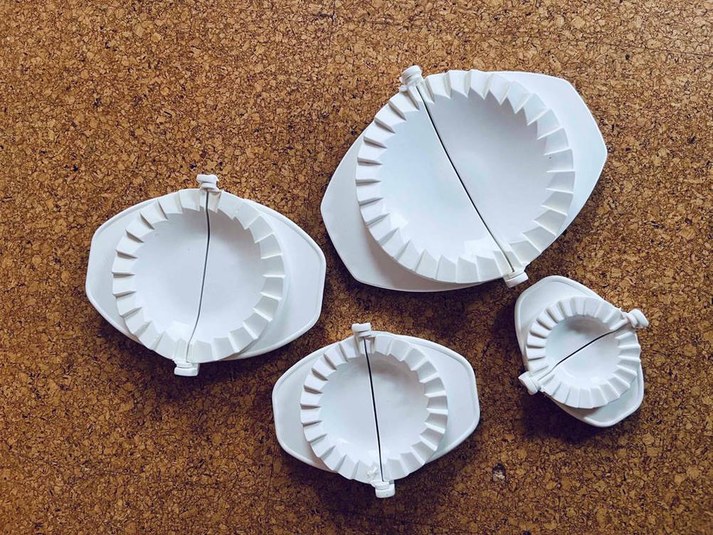 Vier unterschiedlich große, weiße Ravioli-Former im Test liegen geöffnet auf einer Korkunterlage.