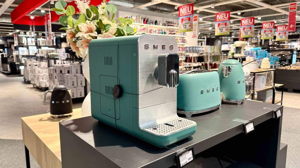 Seitenansicht von schräg vorn des schmalen Kaffeevollautomaten im mintgrünen Design, wie er auf einer Ausstellungsfläche neben weiteren SMEG-Produkten in Mintgrün steht.