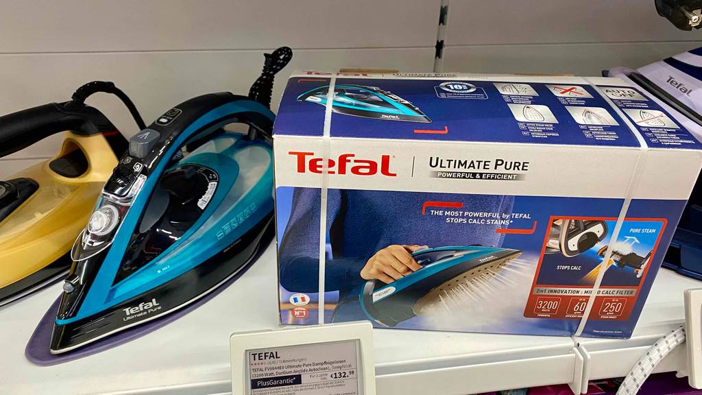 Tefal-Bügeleisen-Test: ein türkis-schwarzes Modell steht neben seiner Originalverpackung auf einem weißen Verkaufsregal zwischen weiteren Bügeleisen.